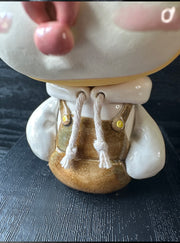 Ceramic sculpture of a large headed character, with closed eyes and full lips, making a kissy face. They have big ears and wear a hoodie, with arms tucked in. Their head is orange with sprinkles atop, like a donut.