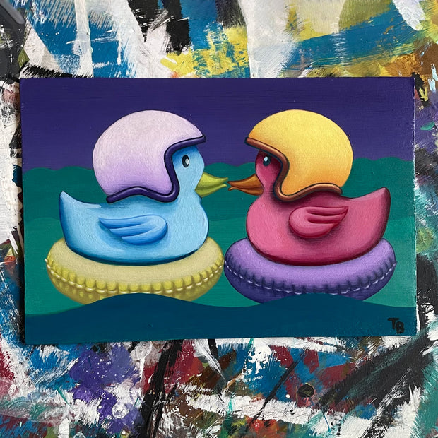 Painting of 2 rubber ducks, one blue and one purple, sitting atop of inflatable pool floaters. They both wear visor-less motorcycle helmets and face one another, with their beaks kissing.