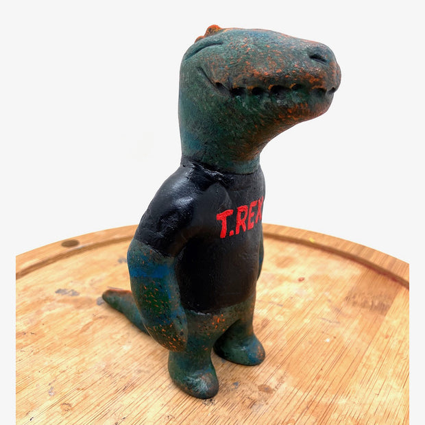 Sculpture of a dark bluish green dinosaur with a simple cartoon face and a wide closed mouth smile and a t-shirt that reads "T-Rex" in red.