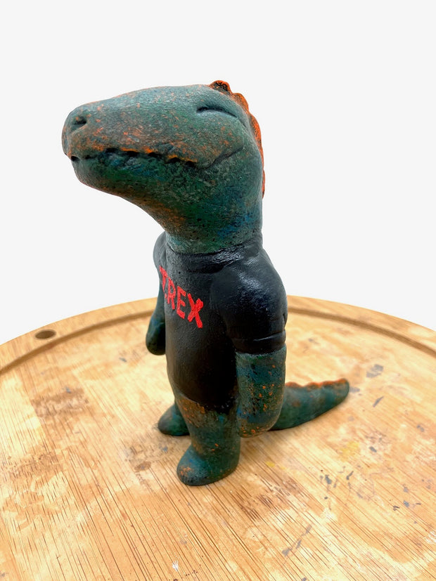 Sculpture of a dark bluish green dinosaur with a simple cartoon face and a wide closed mouth smile and a t-shirt that reads "T-Rex" in red.