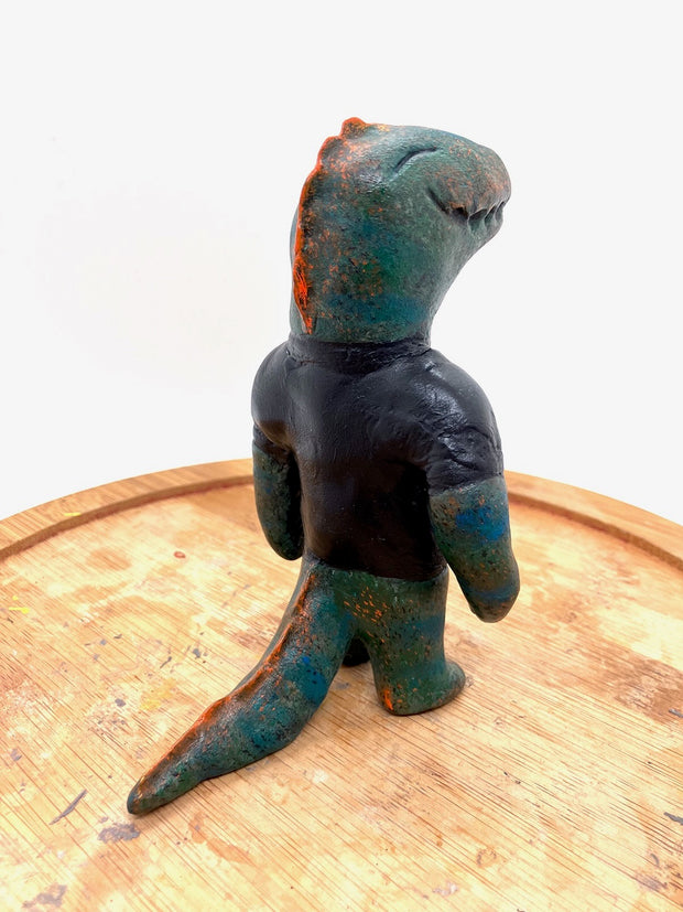 Sculpture of a dark bluish green dinosaur with a simple cartoon face and a wide closed mouth smile and a t-shirt that reads "T-Rex" in red.