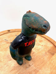 Sculpture of a dark bluish green dinosaur with a simple cartoon face and a wide closed mouth smile and a t-shirt that reads "T-Rex" in red.