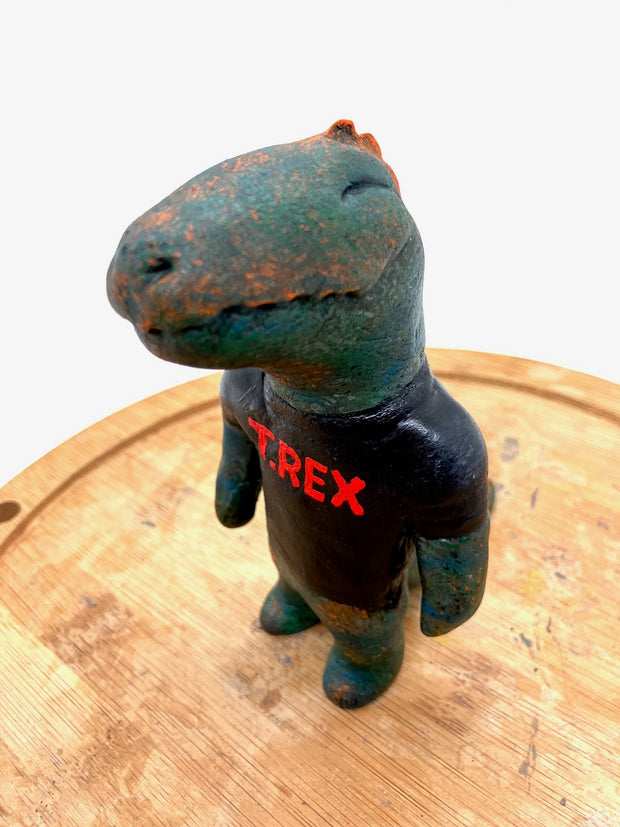 Sculpture of a dark bluish green dinosaur with a simple cartoon face and a wide closed mouth smile and a t-shirt that reads "T-Rex" in red.