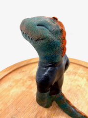 Sculpture of a dark bluish green dinosaur with a simple cartoon face and a wide closed mouth smile and a t-shirt that reads "T-Rex" in red.