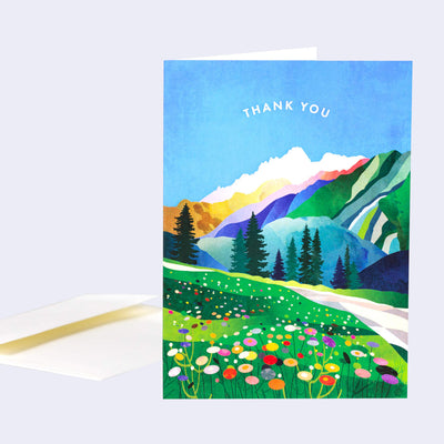 Greeting card with an illustration of a mountain scene with a flowery meadow in the foreground. Text in the blue sky reads "Thank You"