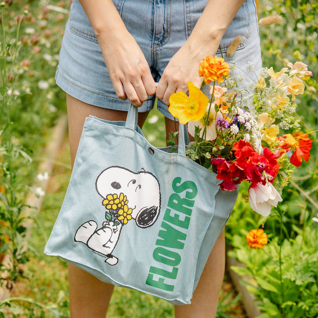 Light blue fabric tote bag with a short purse-like handle and a snap closure. On the front is a graphic of Snoopy, sitting and smiling while holding a yellow bouquet of flowers. Large green text reads "Flowers" along the side.