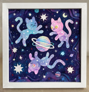 Cut paper artwork of 3 purple, pink and blue mixed colored cats floating around a colorful planet with 2 rings. They all hover amongst many stars on a dark purple background.