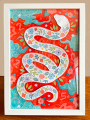 Paper cut art of a white snake with various flowers and leaves on its body in a pattern. Background is bright red with teal blue clouds and cut paper leaves and flowers. Piece is in a white frame.