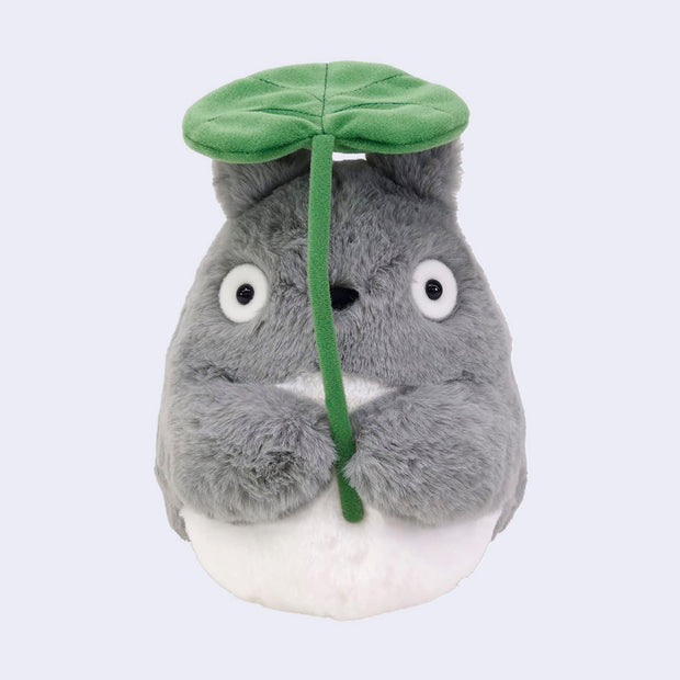 Plush doll of Totoro holding a large green leaf in front of its face and overhead. 