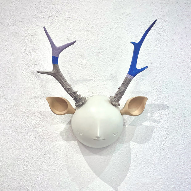 Sculpture of a white round moon-like head with a simple closed eye expression face. It has deer ears and bumpy antlers growing out of its head.