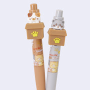 2 retractable pens with cats inside of cardboard boxes as the pen toppers. One cat is gray with a taunting expression and matches its gray pen body, and the other is a sad looking calico cat with a brown pen body.