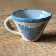 Ceramic mug, mostly cream colored with a blue rim and design of a cat's head on the front of the mug. It smiles with wide eyes and an open outh.