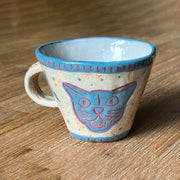 Ceramic mug, mostly cream colored with a blue rim and design of a cat's head on the front, with a wide eye expression and an open mouth smile.