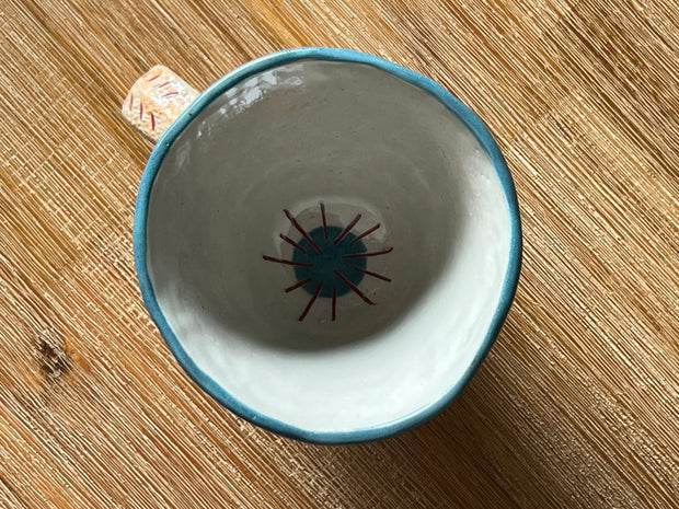 Overhead view of a cream colored ceramic mug with a blue rim and an inner design of a blue circle with red lines coming out from all sides.