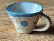 Ceramic mug, mostly cream colored with a blue rim and design of a blue starburst with red lines on the back of the mug.