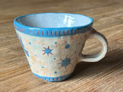 Ceramic mug, mostly cream colored with a blue rim and design of several blue dots with red lines making a starburst design on the back of the mug.