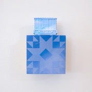 Painted small wooden square panel of a blue sky with a geometric pattern akin to a quilt. Resting atop is a small sculpture of a train car, painting all blue like a sky.