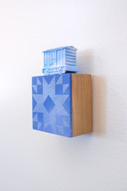 Painted small wooden square panel of a blue sky with a geometric pattern akin to a quilt. Resting atop is a small sculpture of a train car, painting all blue like a sky.