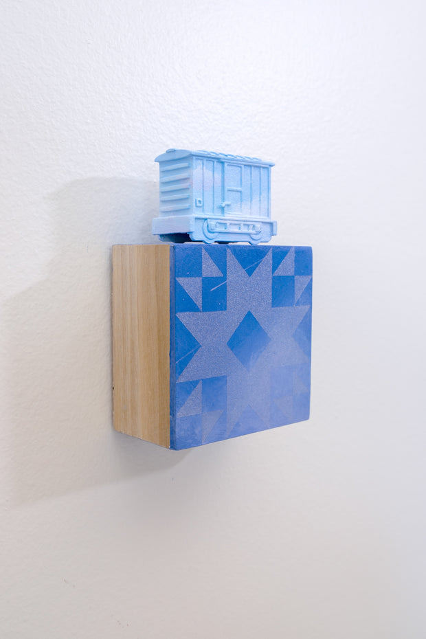 Painted small wooden square panel of a blue sky with a geometric pattern akin to a quilt. Resting atop is a small sculpture of a train car, painting all blue like a sky.