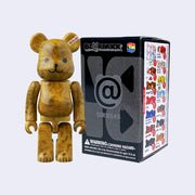 Vinyl bear figure with blocky body features, like an action figure. It has a cute teddy bear smile and is colored a marble caramel brown. It stands next to its product packaging.