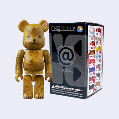 Vinyl bear figure with blocky body features, like an action figure. It has a cute teddy bear smile and is colored a marble caramel brown. It stands next to its product packaging.