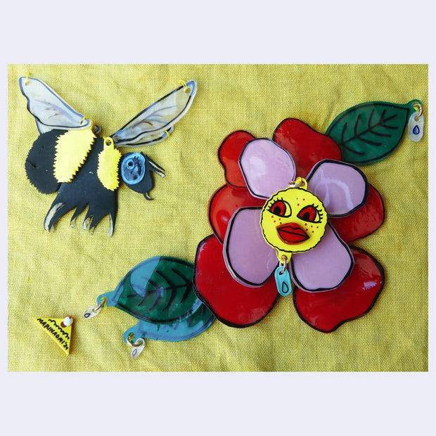 Collage of plastic pieces depicting a scene of a bee flying towards a red and pink flower, with a face in the center. Background is yellow fabric.