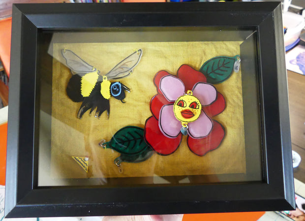 Collage of plastic pieces depicting a scene of a bee flying towards a red and pink flower, with a face in the center. Background is yellow fabric. Piece is in shadowbox frame.