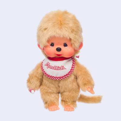 Plush doll of Monchhichi, a cute monkey that has a baby animal face with rosy cheeks and brown eyes. It has beige fluffy fur, wears a white and red plaid bib and has a pacifier in its hand that can go in its mouth.