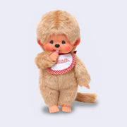 Plush doll of Monchhichi, a cute monkey that has a baby animal face with rosy cheeks and brown eyes. It has beige fluffy fur, wears a white and red plaid bib and has a pacifier in its hand that can go in its mouth.