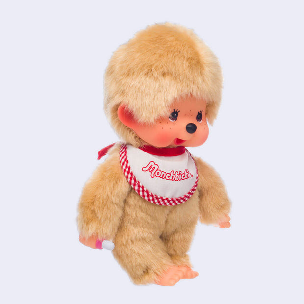 Plush doll of Monchhichi, a cute monkey that has a baby animal face with rosy cheeks and brown eyes. It has beige fluffy fur, wears a white and red plaid bib and has a pacifier in its hand that can go in its mouth.