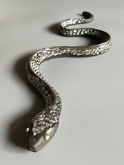 Ceramic sculpture of a snake, with a long dark silver body and silver eyes. It is seen from a front view, slithering forward.