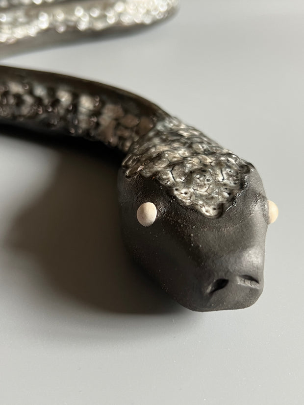 Ceramic sculpture of a snake, with a long dark silver body and silver eyes. 