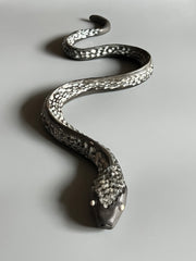 Ceramic sculpture of a snake, with a long dark silver body and silver eyes. It is seen from overhead, slithering forward.