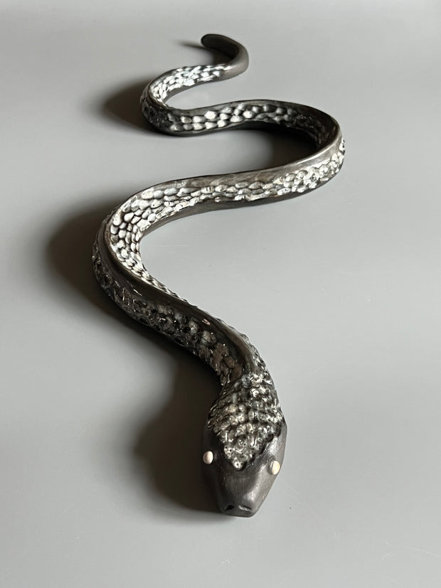 Ceramic sculpture of a snake, with a long dark silver body and silver eyes. It is seen from overhead, slithering forward.