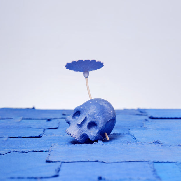 Assembled carved wooden sculpture of a purple skull with a stemmed flower growing out of its head, with the other end of the stem visible from the opposite side of the skull.