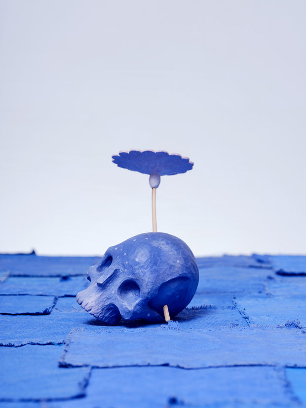 Assembled carved wooden sculpture of a purple skull with a stemmed flower growing out of its head, with the other end of the stem visible from the opposite side of the skull.