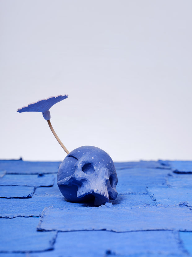 Assembled carved wooden sculpture of a purple skull with a stemmed flower growing out of its head, with the other end of the stem visible from the opposite side of the skull.