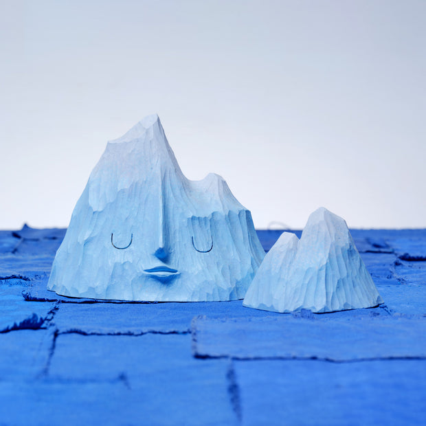 Set of 2 carved wooden sculptures, painting blue, of mountain ranges. One has a closed eye, calm facial expression and the other is without a face and smaller in size.