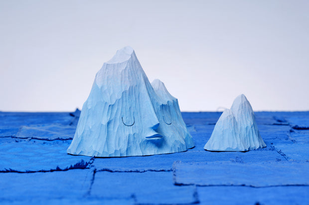 Set of 2 carved wooden sculptures, painting blue, of mountain ranges. One has a closed eye, calm facial expression and the other is without a face and smaller in size.