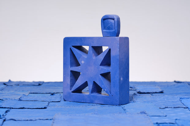 Carved wooden sculpture made to look like a cement breeze block with a centralized star pattern. The block is entirely purplish blue and a small TV sits on top of it.