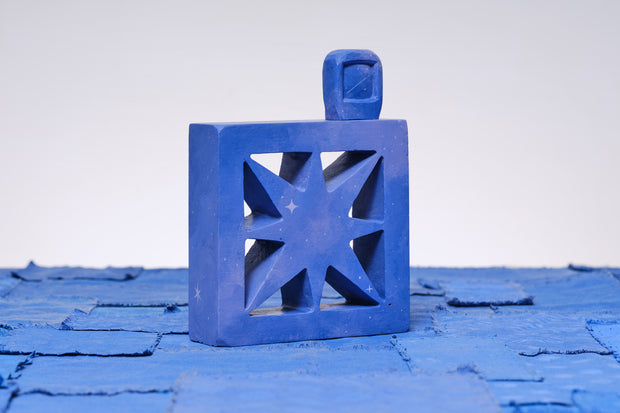 Carved wooden sculpture made to look like a cement breeze block with a centralized star pattern. The block is entirely purplish blue and a small TV sits on top of it.