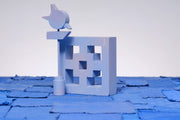 Carved wooden sculpture made to look like a cement breeze block. The block is entirely painted a light purplish blue and a tall sign post stands nearby, painted in similar colors.