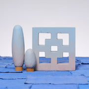 Carved wooden sculpture made to look like a cement breeze block. The block is entirely painted a light blue to pink gradient. Two oval shaped trees of varying heights stand nearby, painted in similar color tones.