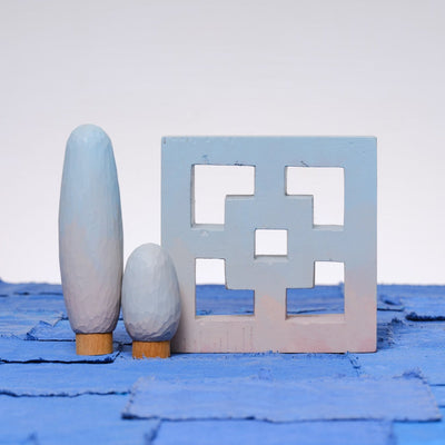 Carved wooden sculpture made to look like a cement breeze block. The block is entirely painted a light blue to pink gradient. Two oval shaped trees of varying heights stand nearby, painted in similar color tones.