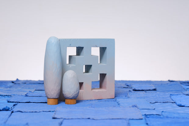 Carved wooden sculpture made to look like a cement breeze block. The block is entirely painted a light blue to pink gradient. Two oval shaped trees of varying heights stand nearby, painted in similar color tones.