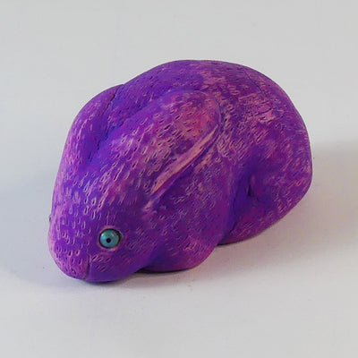 Sculpture of a pinkish-purple bunny with blue eyes. It sits huddled into itself, without any limbs exposed.