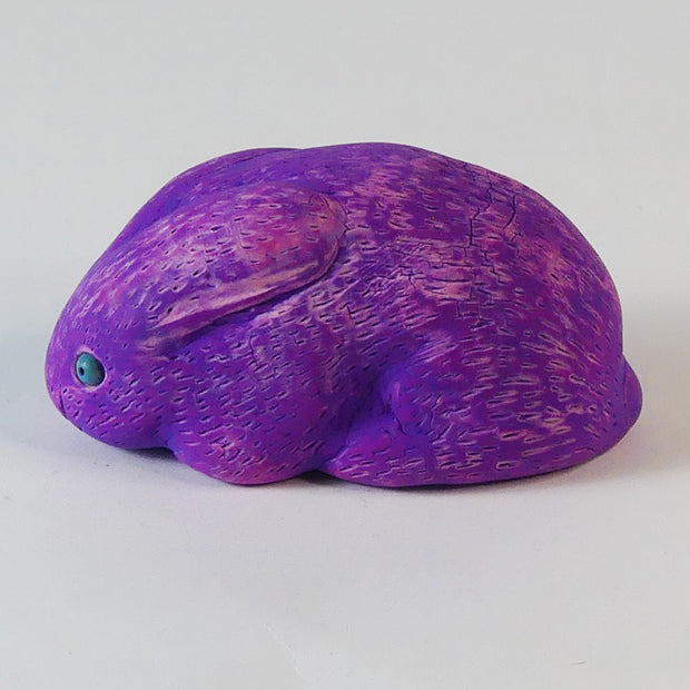 Sculpture of a pinkish-purple bunny with blue eyes. It sits huddled into itself, without any limbs exposed.