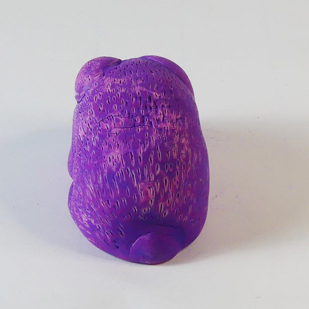 Sculpture of a pinkish-purple bunny with blue eyes. It sits huddled into itself, without any limbs exposed.