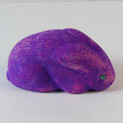 Sculpture of a pinkish-purple bunny with blue eyes. It sits huddled into itself, without any limbs exposed.