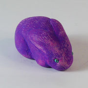 Sculpture of a pinkish-purple bunny with blue eyes. It sits huddled into itself, without any limbs exposed.